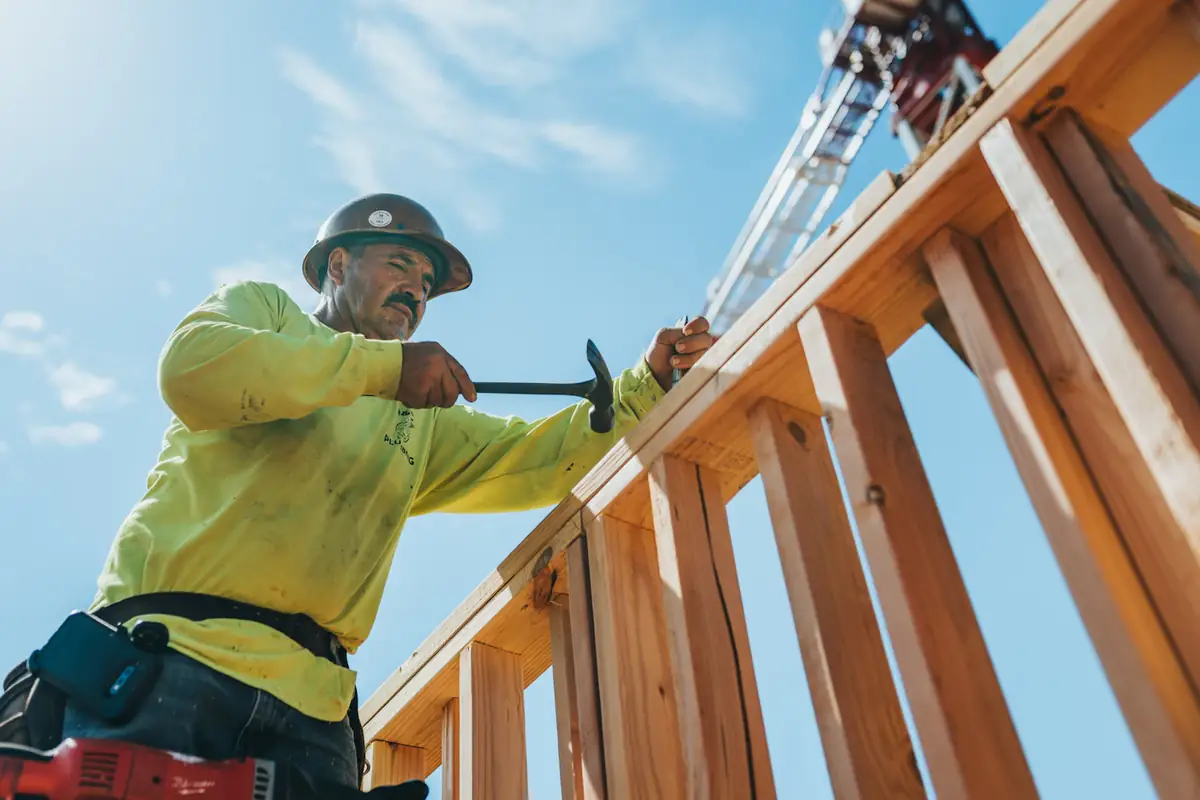 A builder in a hard hat nailing up a timber frame against a blue sky