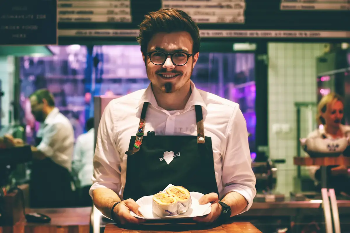 A restaurant owner in an apron holding a plate in a busy dining room