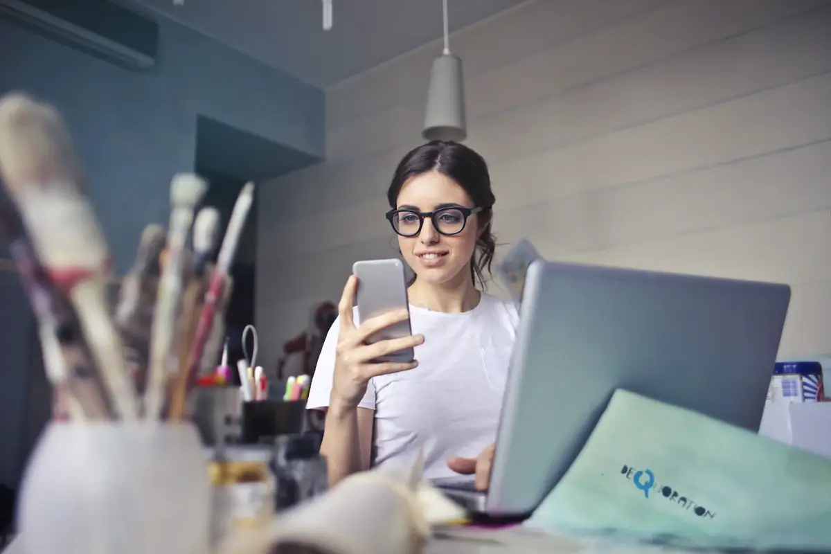 A woman checking her phone at her desk beside a laptop