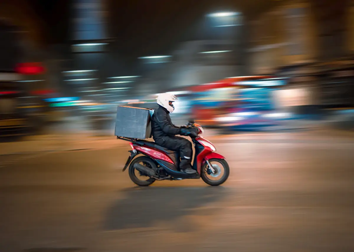 A delivery rider on a scooter at night, lights streaking past