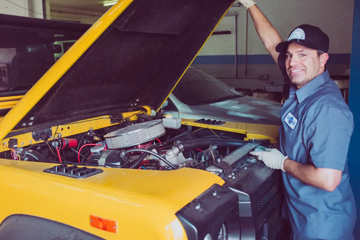 A mechanic smiling under the open bonnet of a yellow car